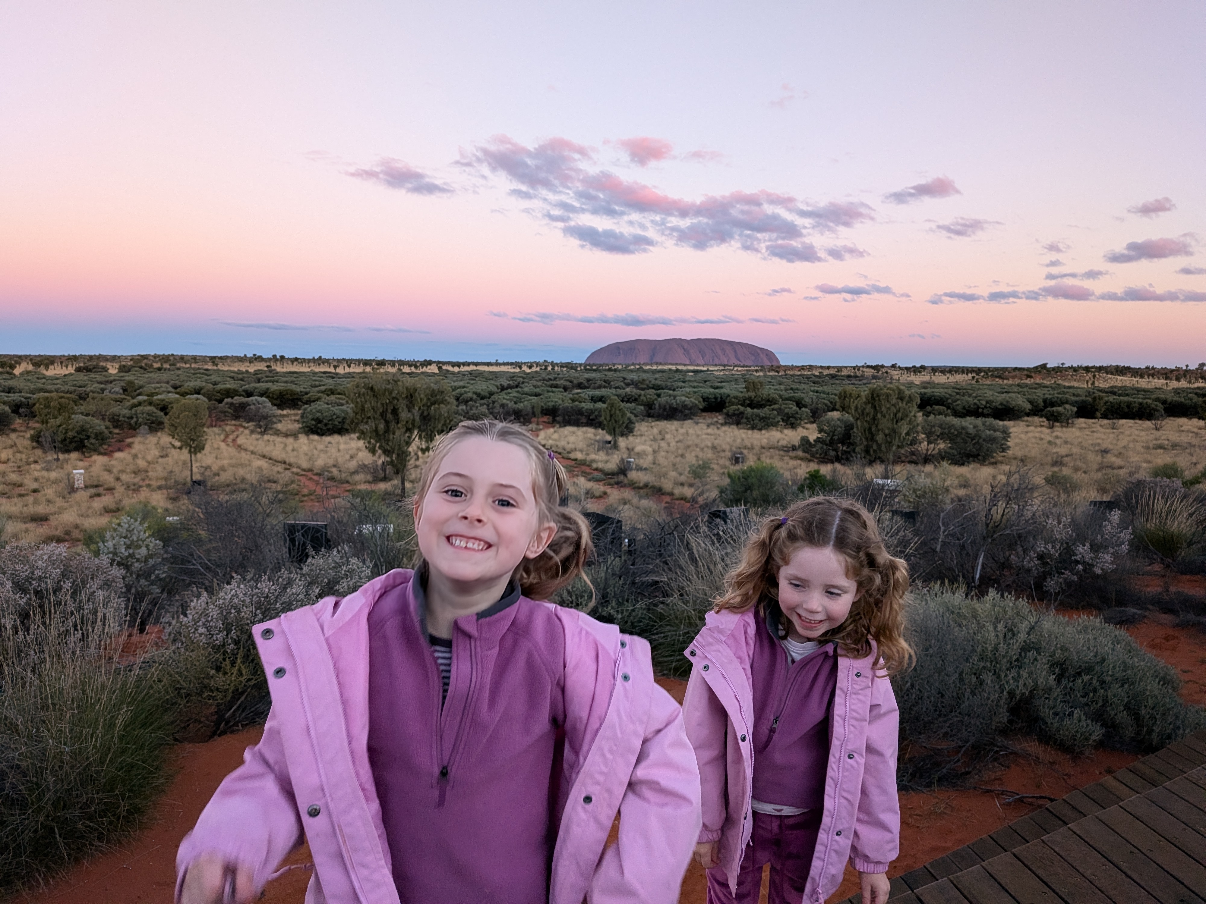 The girls grinning with pink sunset sky and Uluru behind at Wintjiri Wiru