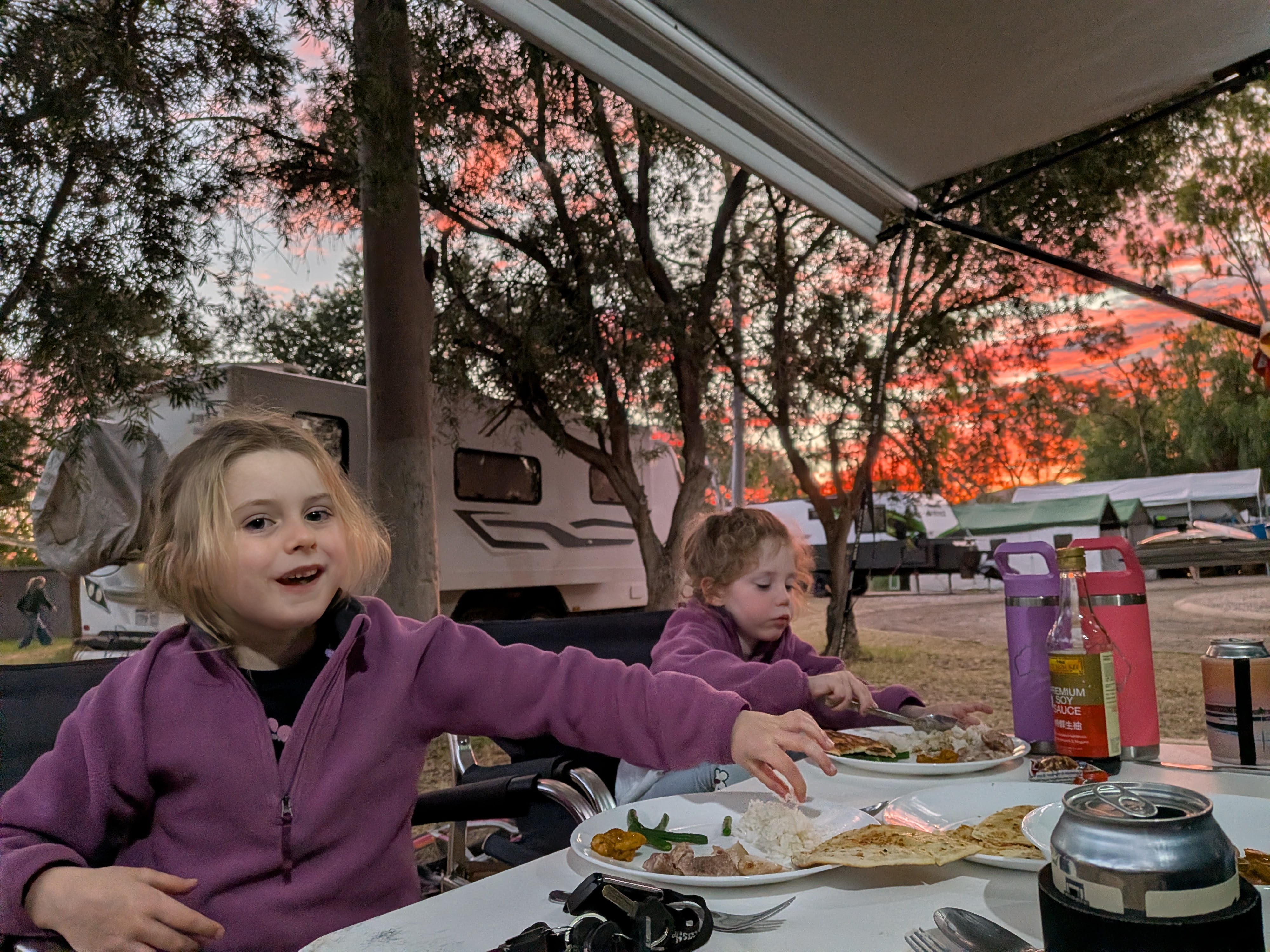 The girls eating butter chicken and naan at the camp table under a pink sunset