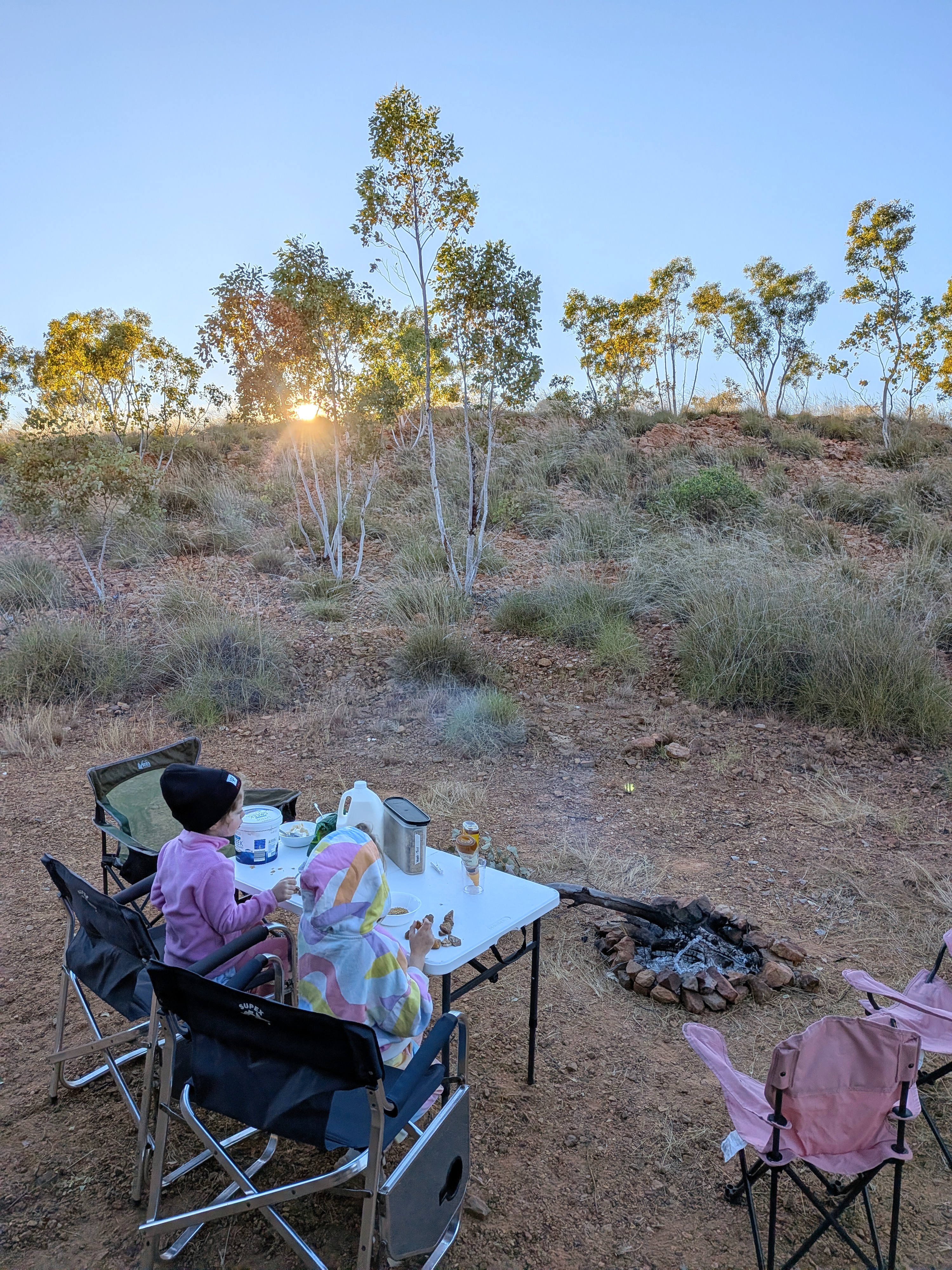The girls eating breakfast at the camp table by the fire as the sun rises over the ridge at Churchill's Head