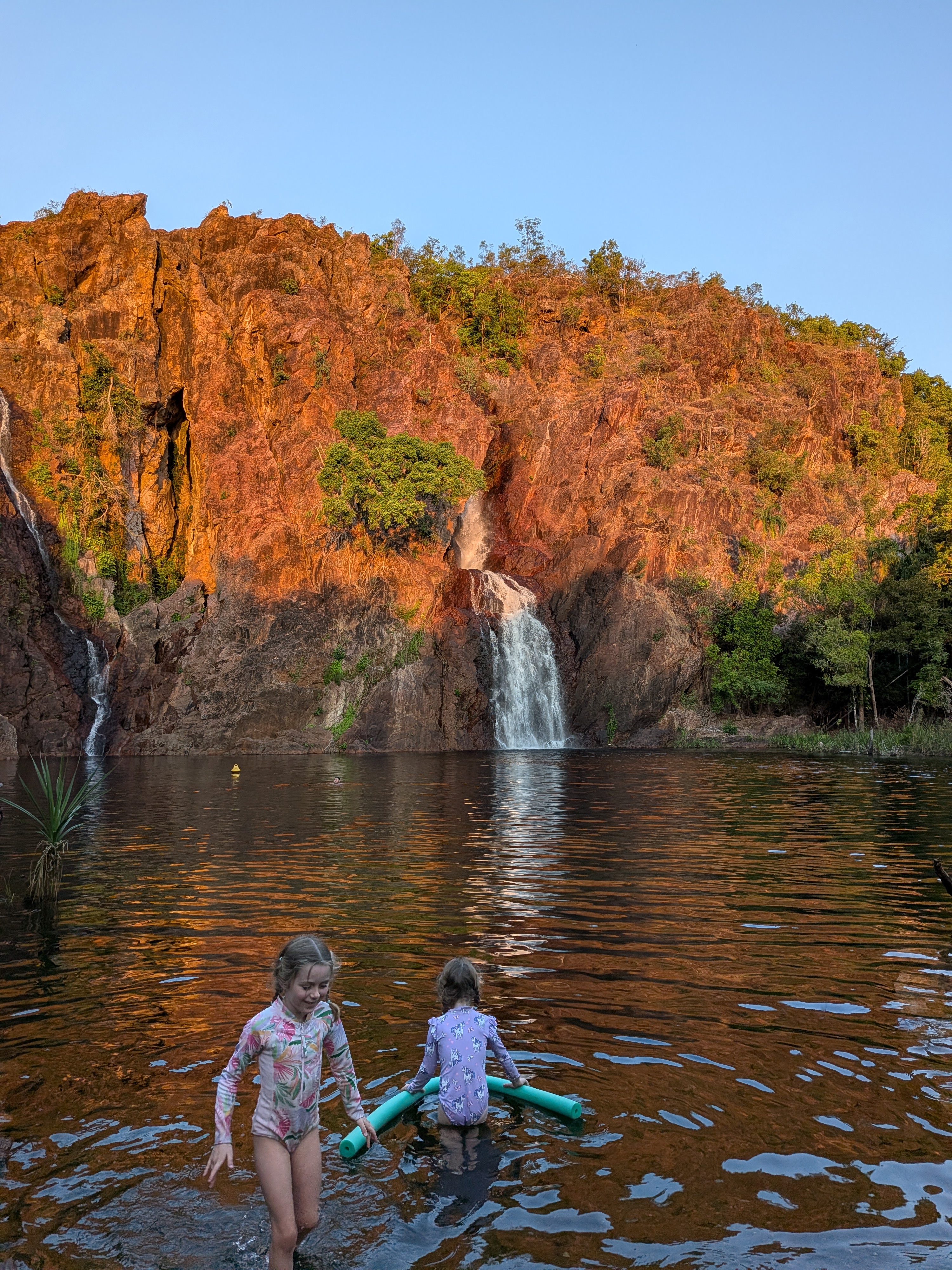 The girls in the shallows of the Wangi Falls plunge pool as the cliff glows red at sunset