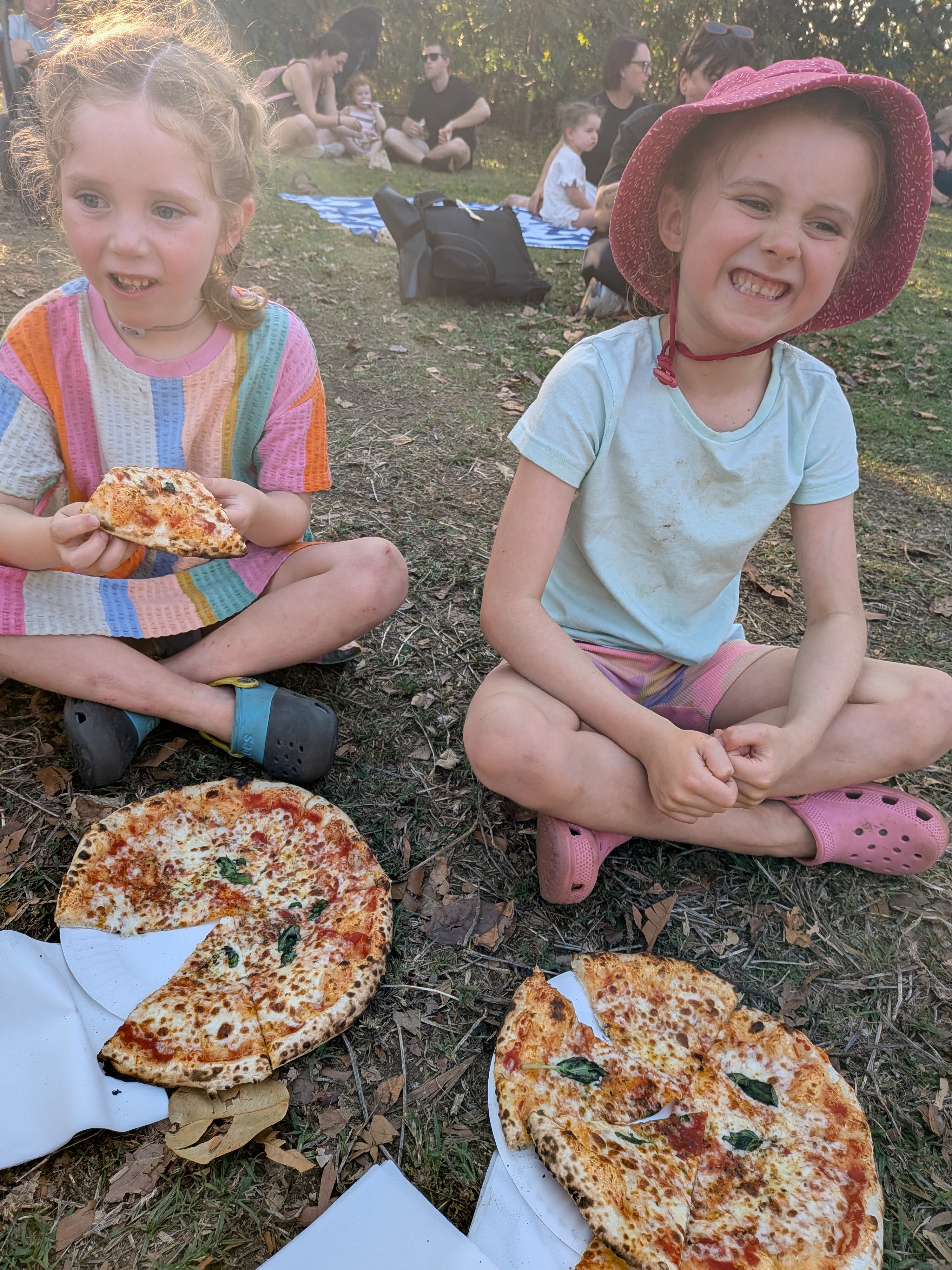Leah and Hazel sitting cross-legged on the grass with a margherita pizza each at the markets