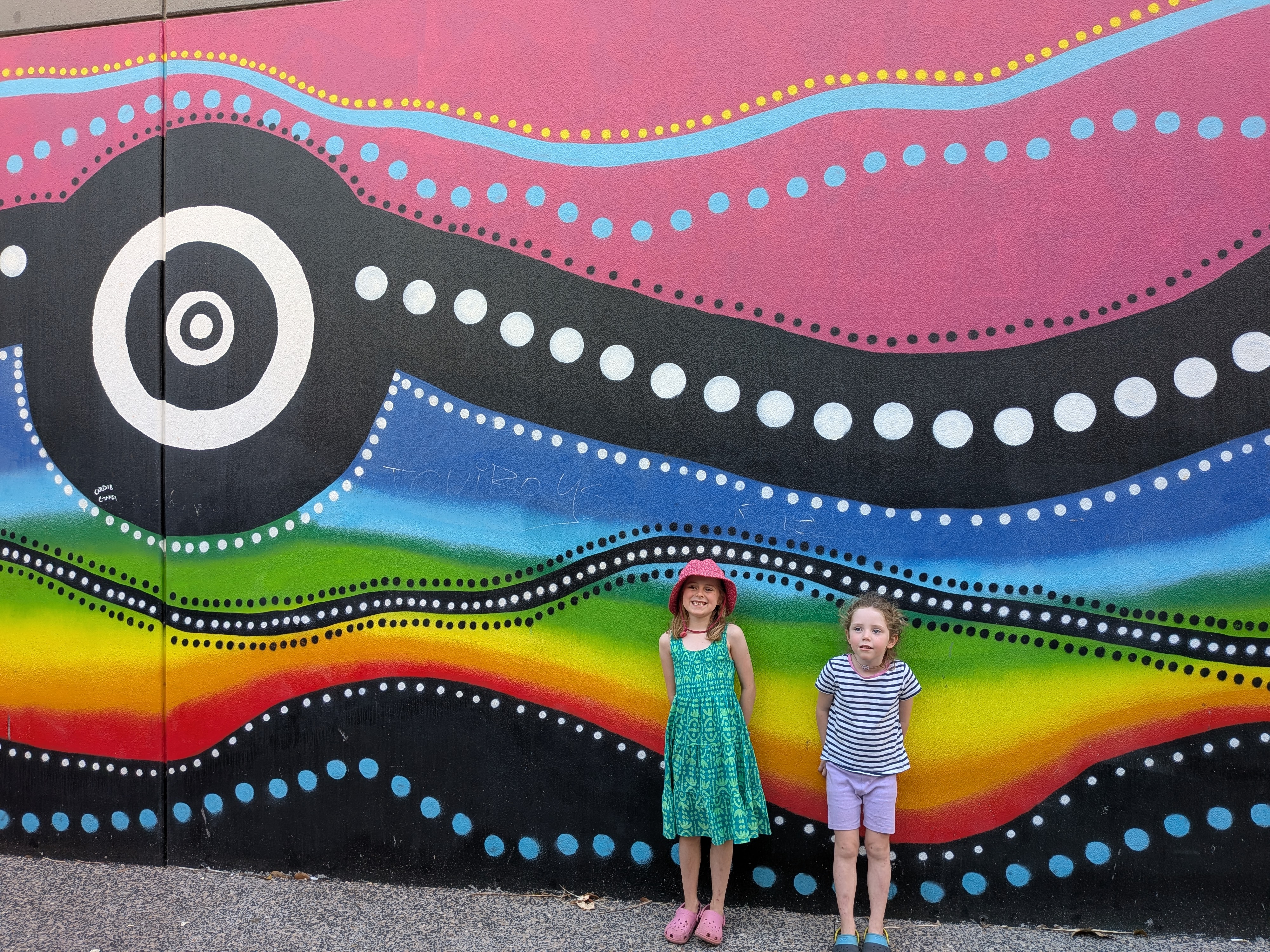 Hazel and Leah in front of a huge dot-painted rainbow mural in the city