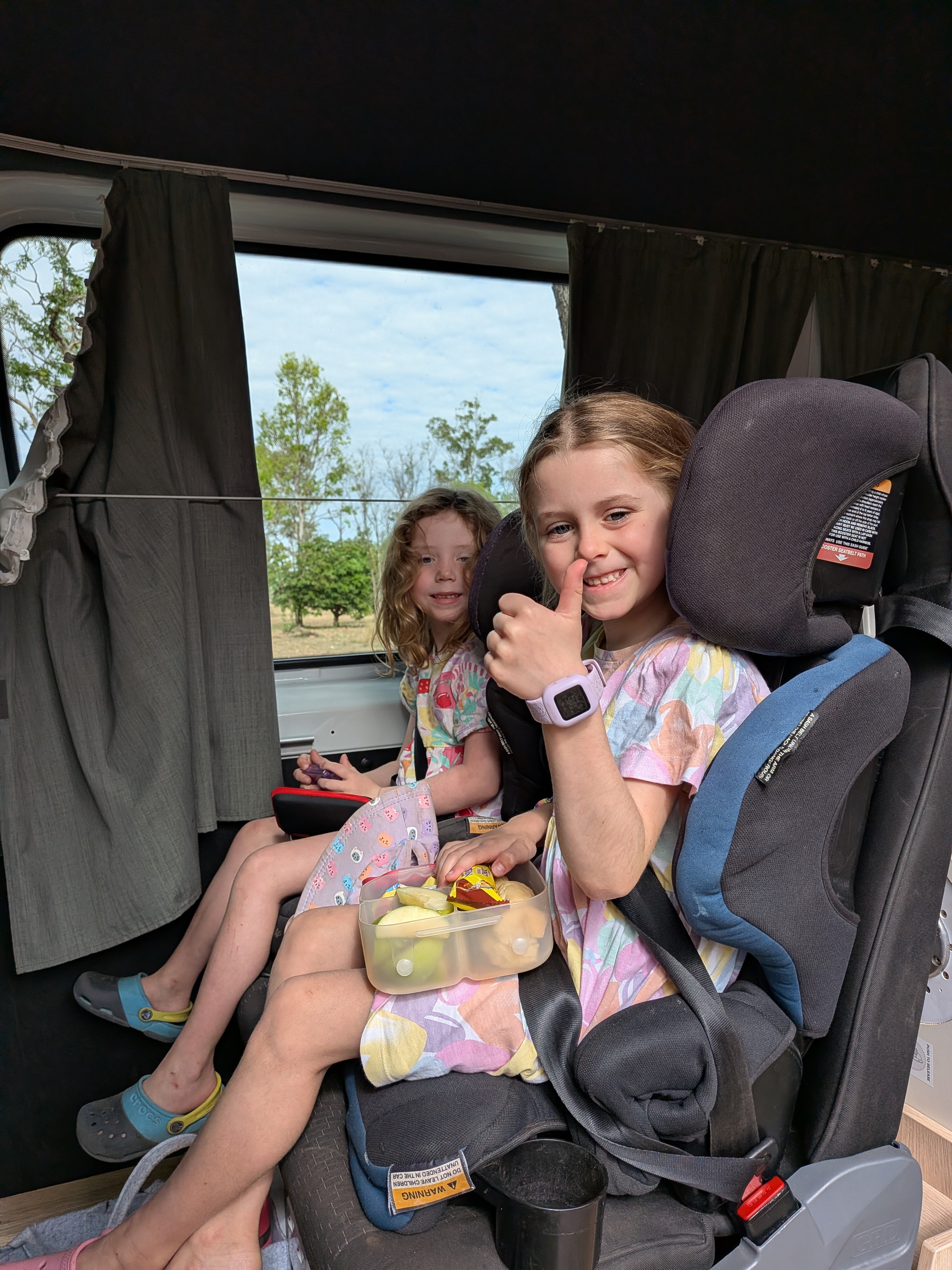Hazel and Leah buckled into Lizzie&rsquo;s car seats, snacks and thumbs at the ready