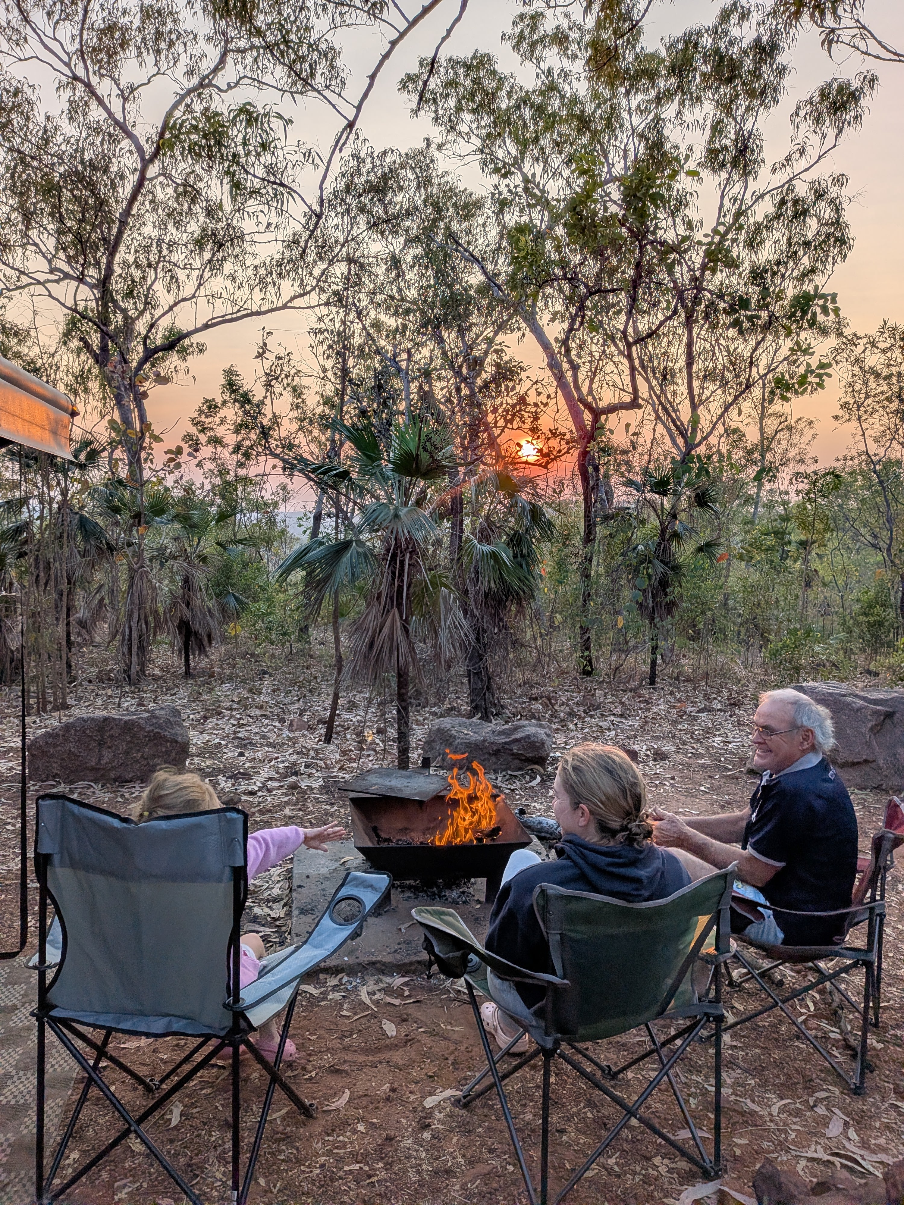 Three around the morning fire as the sun comes up through the trees