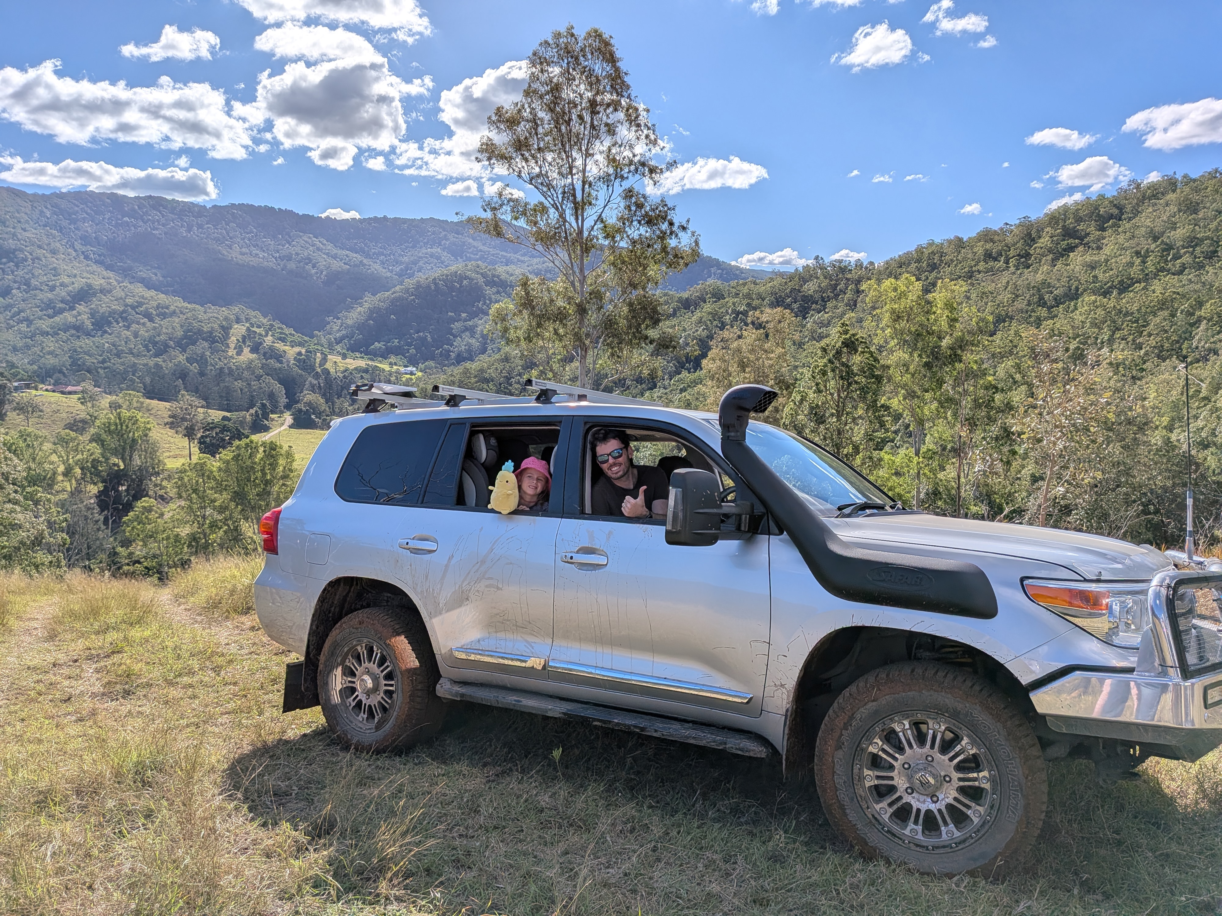 Reg the silver Landcruiser on a grassy hilltop with Dylan giving a thumbs up out the window and Leah in the back seat