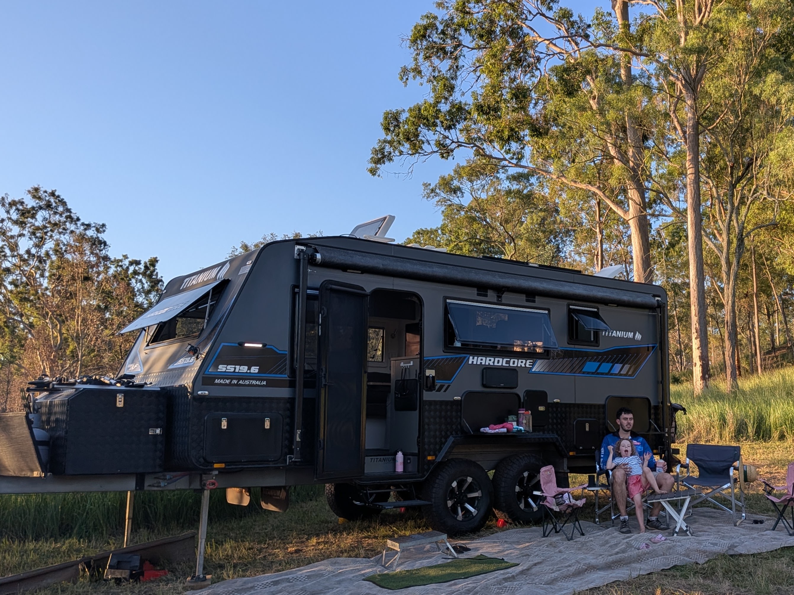 The TITANIUM HARDCORE caravan set up at camp among gum trees in the late afternoon sun, with Dylan and Leah relaxing beside it